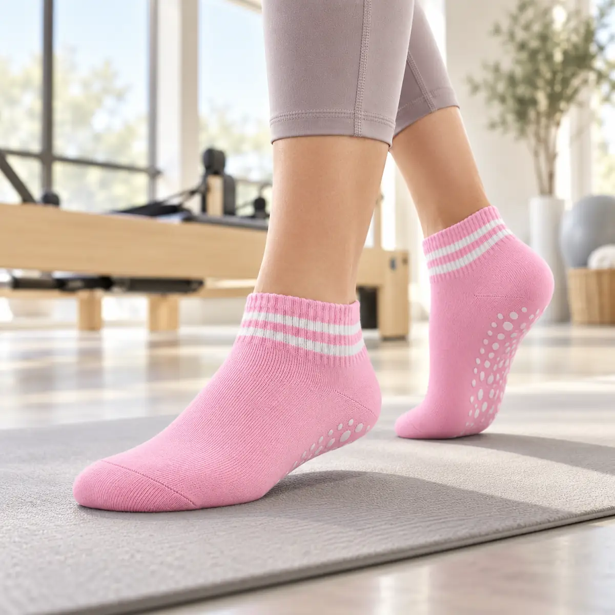 Foto 1 van 3 Roze Enkel sokken met twee witte strepen aan de voeten van een vrouw op een grijze mat, witte noppen onder de opgetilde hiel, lichte studio met een houten reformer onscherp op de achtergrond
